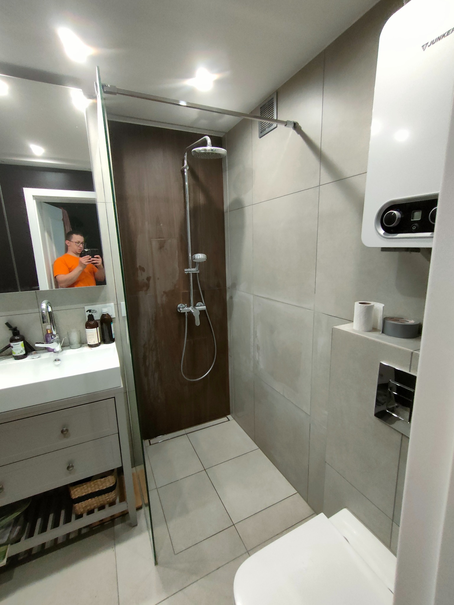 Nowoczesna łazienka z kabiną prysznicową typu walk-in, umywalką z szafką i grzejnikiem Junkers. W lustrze odbija się osoba robiąca zdjęcie.