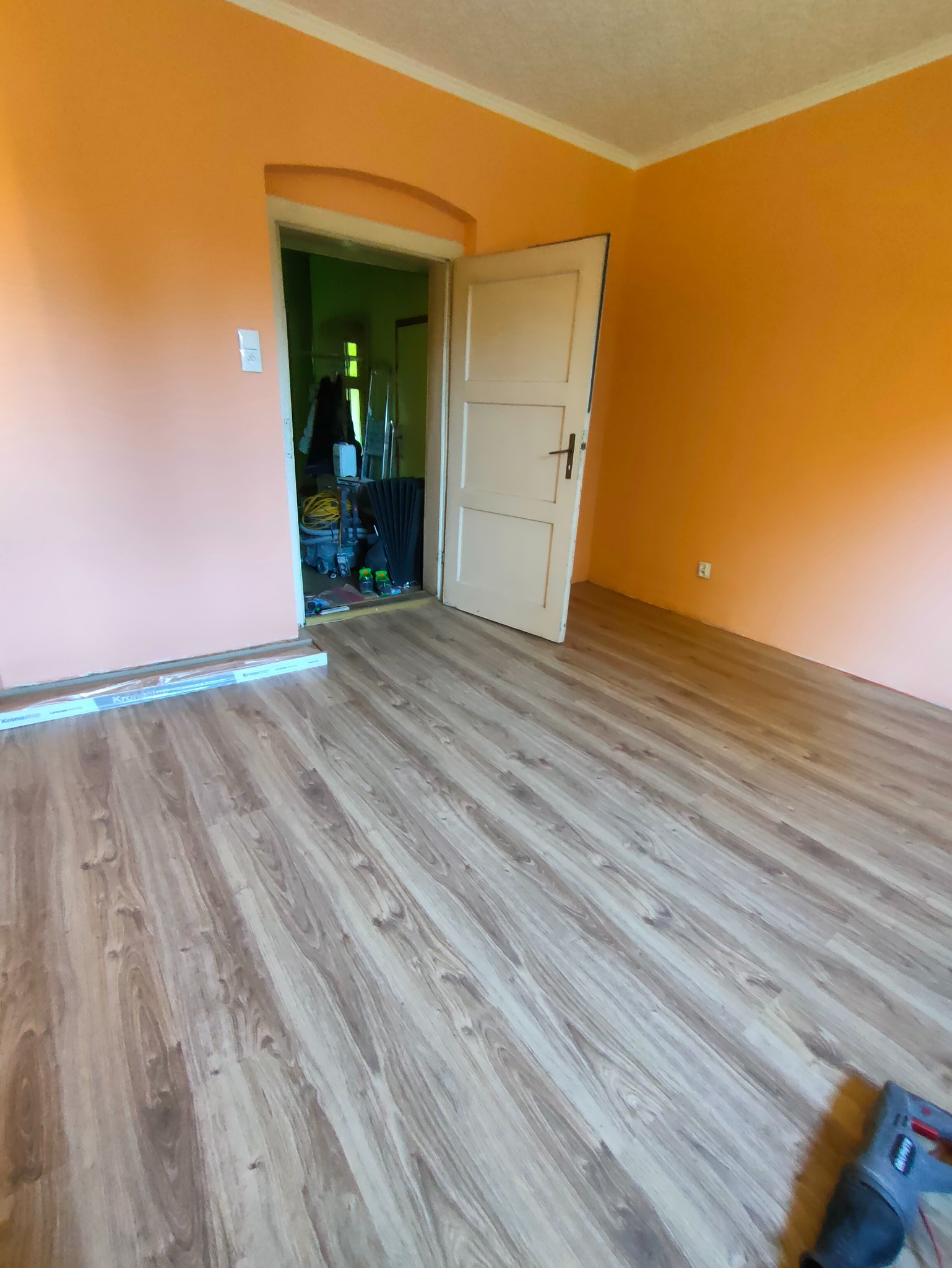 Ułożone panele podłogowe w pokoju z pomarańczowymi ścianami. Widoczny fragment narzędzia do cięcia paneli i opakowanie paneli przy ścianie. Widok na otwarte drzwi do innego pomieszczenia.