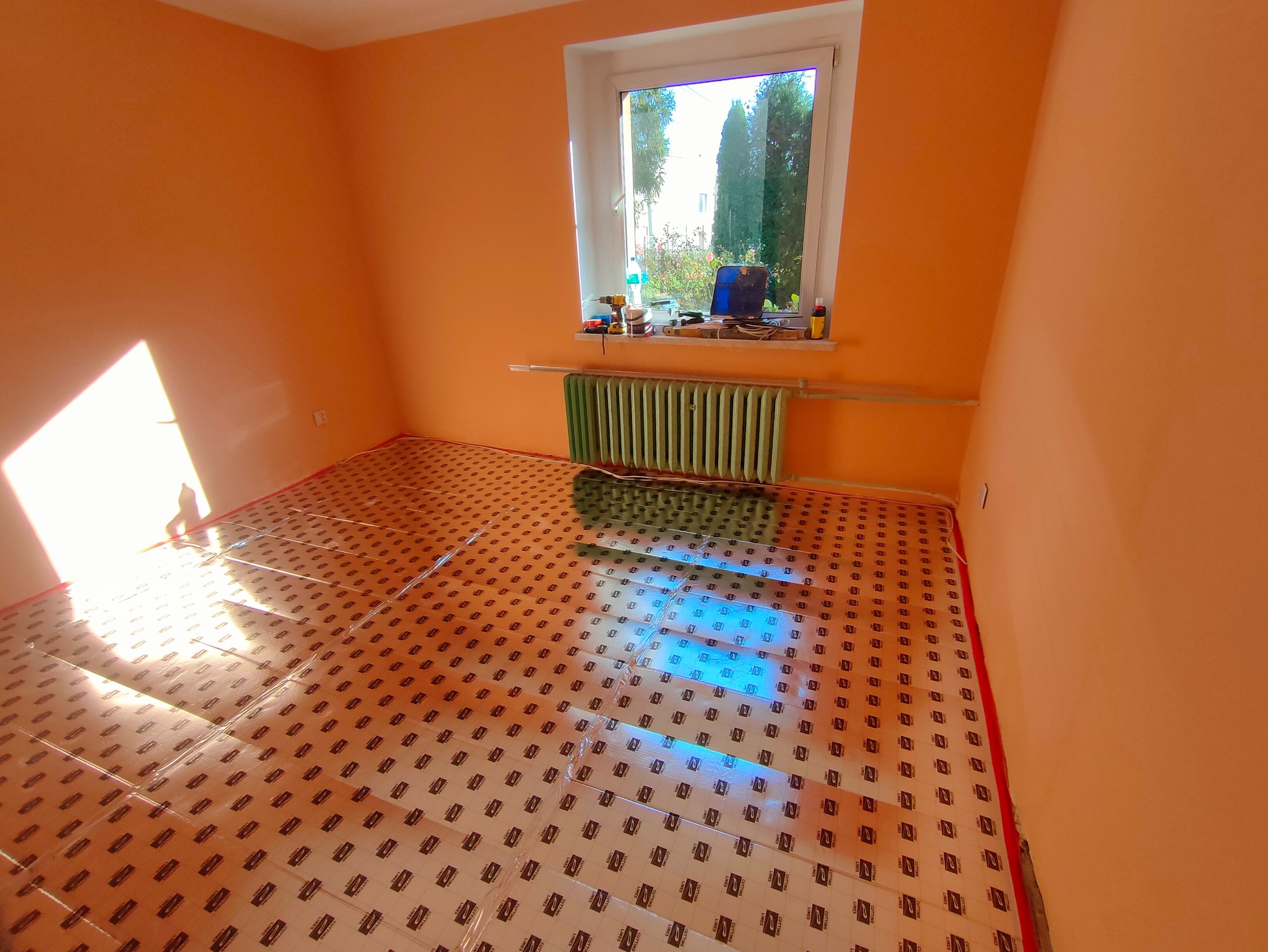 Pomarańczowy pokój z rozłożoną folią grzewczą na podłodze, przygotowaną do dalszych prac. Widoczne narzędzia na parapecie i grzejnik pod oknem.
