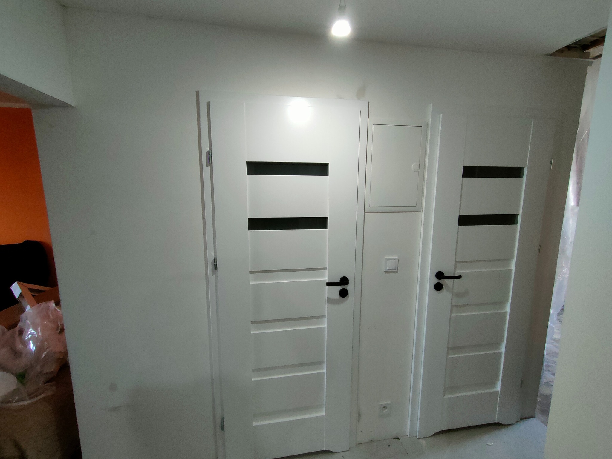 Dwa białe, nowoczesne drzwi wewnętrzne z czarnymi poziomymi wstawkami i klamkami, zamontowane w jasnym pomieszczeniu w trakcie wykańczania.