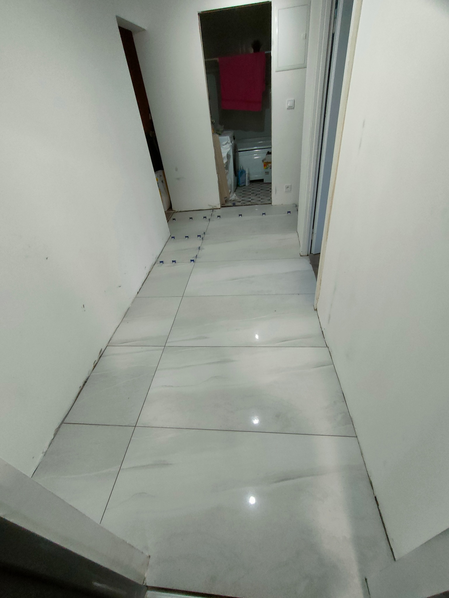 Jasne, duże płytki podłogowe w korytarzu, częściowo ułożone, z niebieskimi krzyżykami dystansowymi. W tle widoczne wejście do łazienki z pralką.