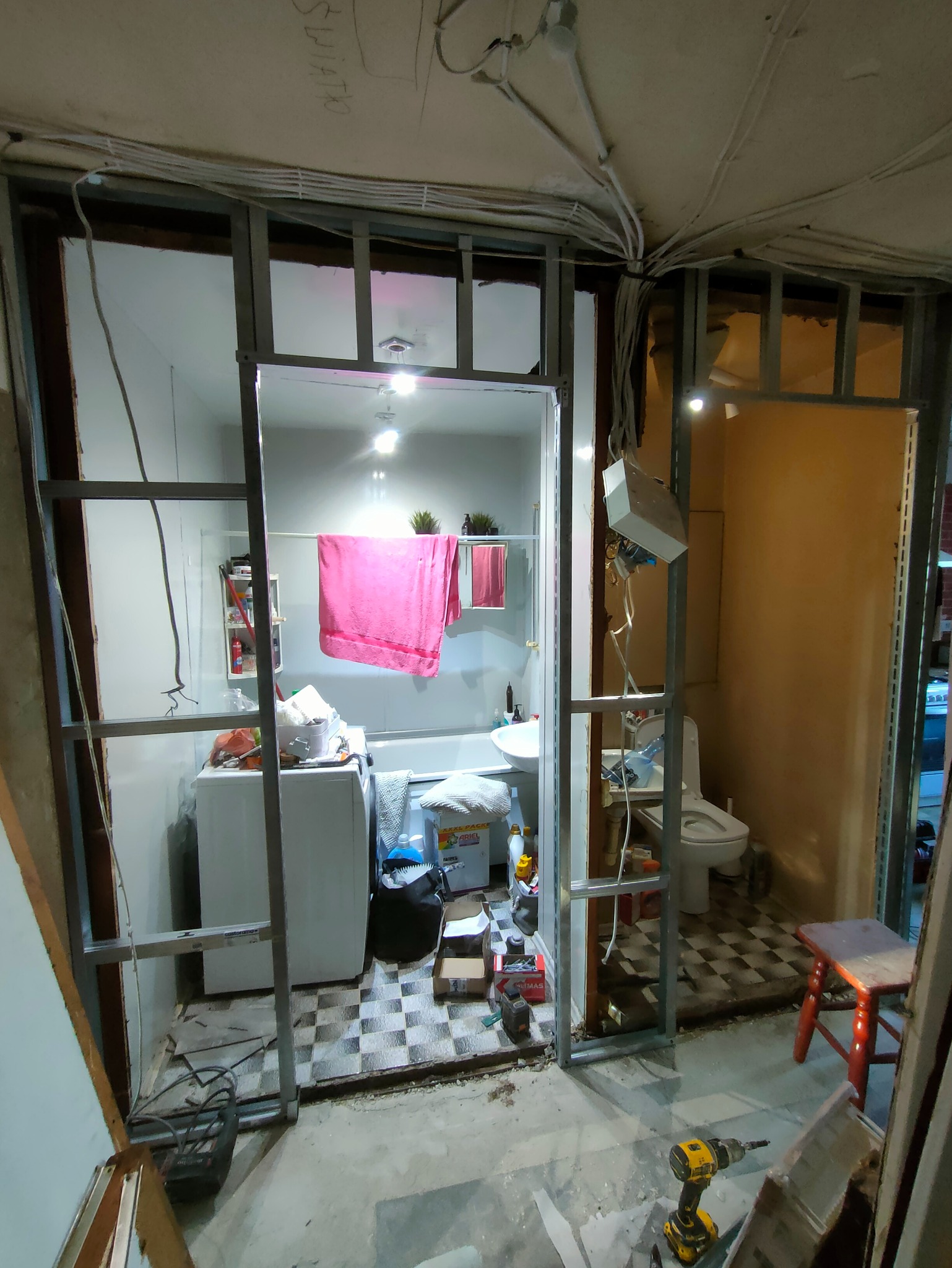 Wnętrze łazienki w trakcie remontu. Widoczne metalowe profile konstrukcyjne, pralka, wanna, toaleta, narzędzia i materiały budowlane. Uwagę zwracają kable elektryczne pod sufitem.