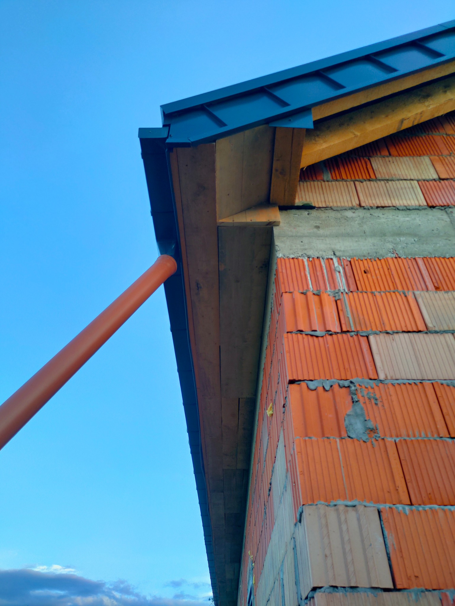 Niski kąt: fragment ściany z czerwonej cegły ceramicznej, z zamontowaną rynną i rurą spustową na tle błękitnego nieba. Widoczny fragment dachu.