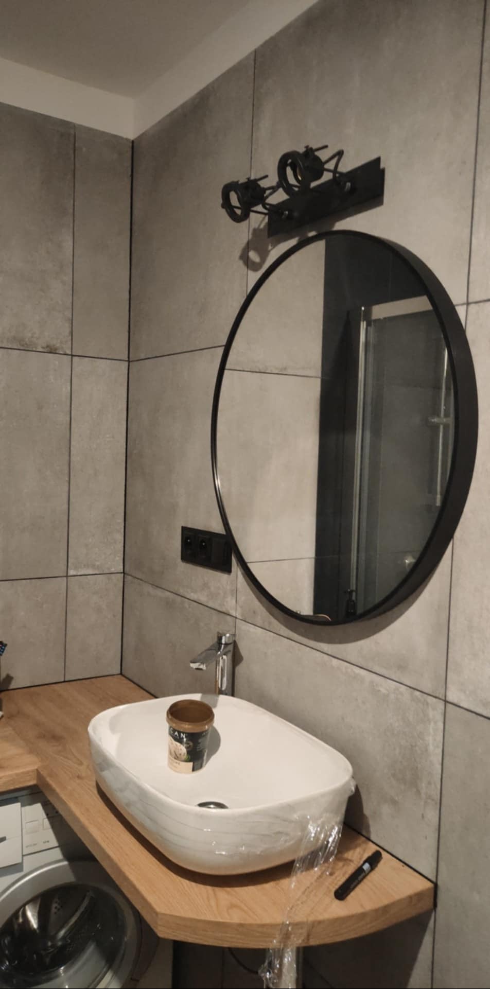 Nowoczesna łazienka z okrągłym lustrem, umywalką na drewnianym blacie i szarą płytką. Widoczna lampa nad lustrem i fragment pralki. Umywalka zabezpieczona folią.