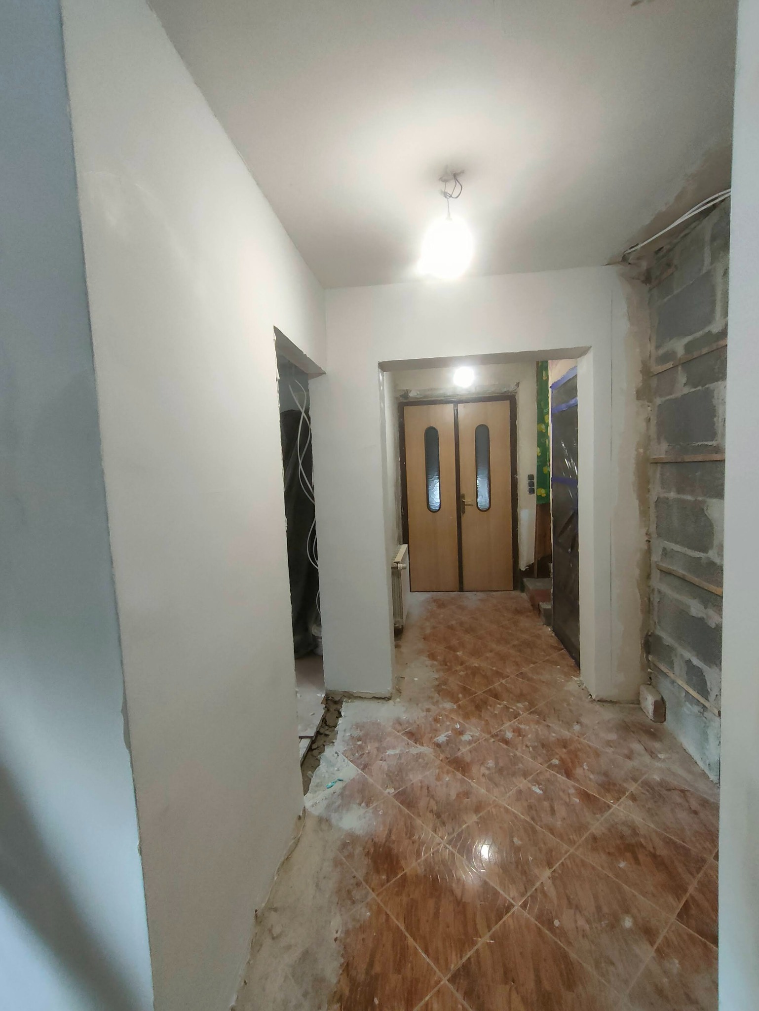 Korytarz w trakcie remontu: częściowo otynkowana ściana, odsłonięte bloczki, stara terakota. Widoczne kable i przygotowania do dalszych prac wykończeniowych.