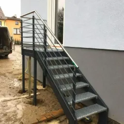 Zewnętrzne schody metalowe z kratowymi stopniami i nierdzewną balustradą, w trakcie montażu; widoczne podpory tymczasowe i przygotowany grunt.