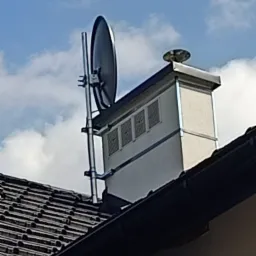 Antena satelitarna zamontowana na kominie wentylacyjnym na dachu budynku, widok z dołu na tle błękitnego nieba z chmurami.