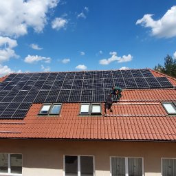 IDEA PV Sp. z o.o. - Montaż paneli fotowoltaicznych na dachu domu z czerwonej dachówki, widoczni dwaj instalatorzy w trakcie pracy, drabina oparta o budynek, błękitne niebo z chmurami w tle.