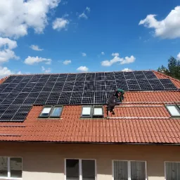Montaż paneli fotowoltaicznych na dachu domu z czerwonej dachówki, widoczni dwaj instalatorzy w trakcie pracy, drabina oparta o budynek, błękitne niebo z chmurami w tle.