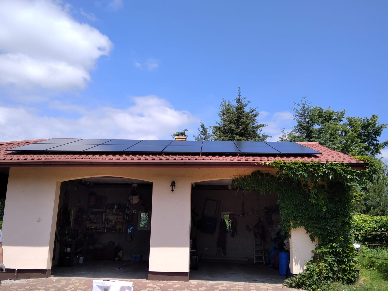 Panele słoneczne na dachu garażu z czerwoną dachówką, widok z dołu, zarośnięty bluszczem fragment budynku.