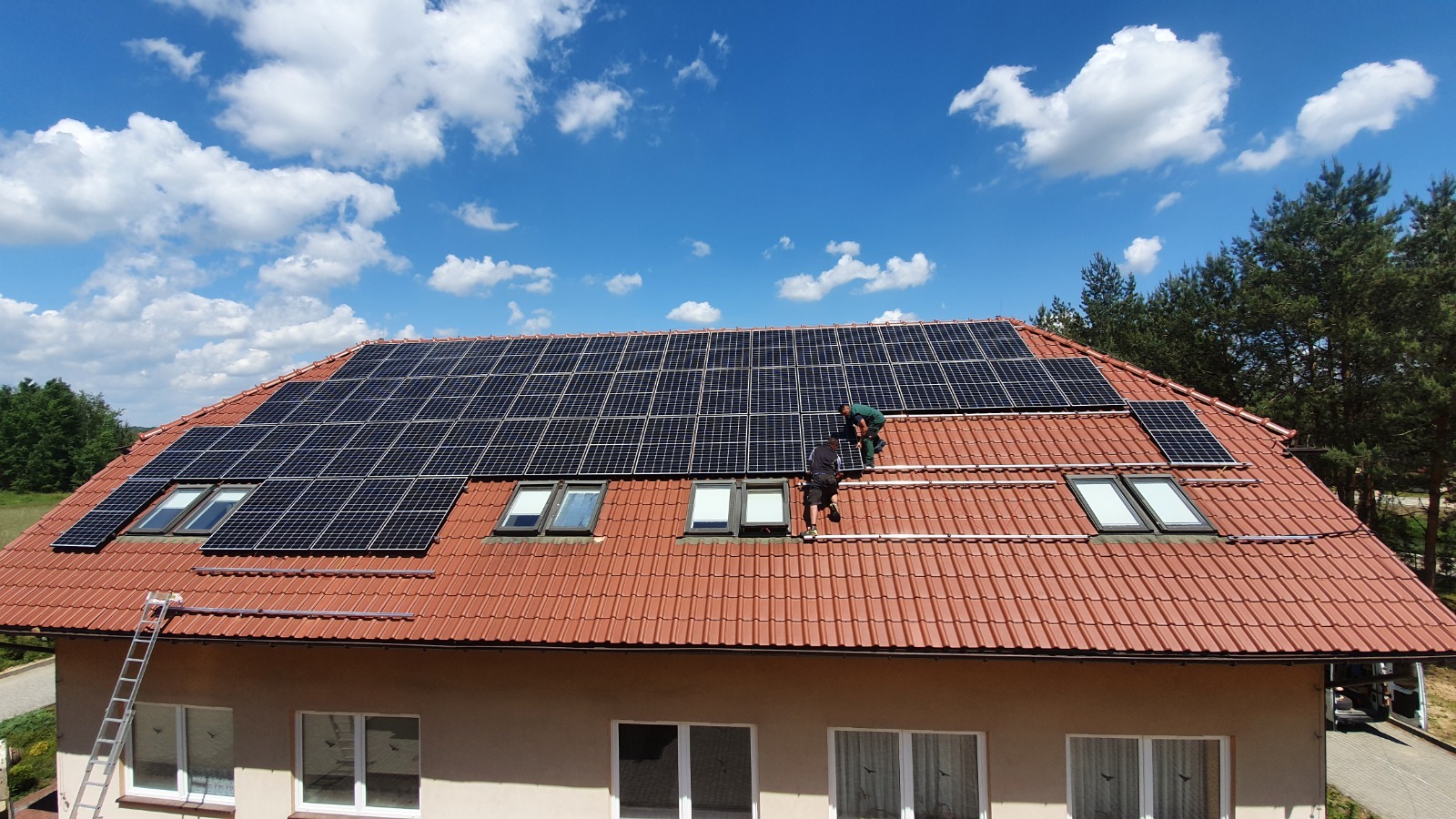 Montaż paneli fotowoltaicznych na dachu domu z czerwonej dachówki, widoczni dwaj instalatorzy w trakcie pracy, drabina oparta o budynek, błękitne niebo z chmurami w tle.