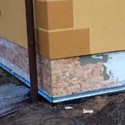 Fragment elewacji budynku z częściowo wykończonym kamiennym cokołem, widoczna rynna spustowa i fragment terenu z ziemią.