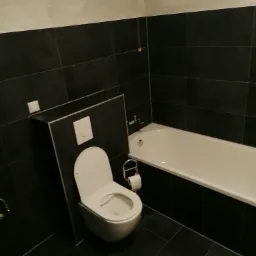 Nowoczesna łazienka z czarnymi płytkami, białą wanną i podwieszaną toaletą z zabudowaną spłuczką, minimalistyczny design.