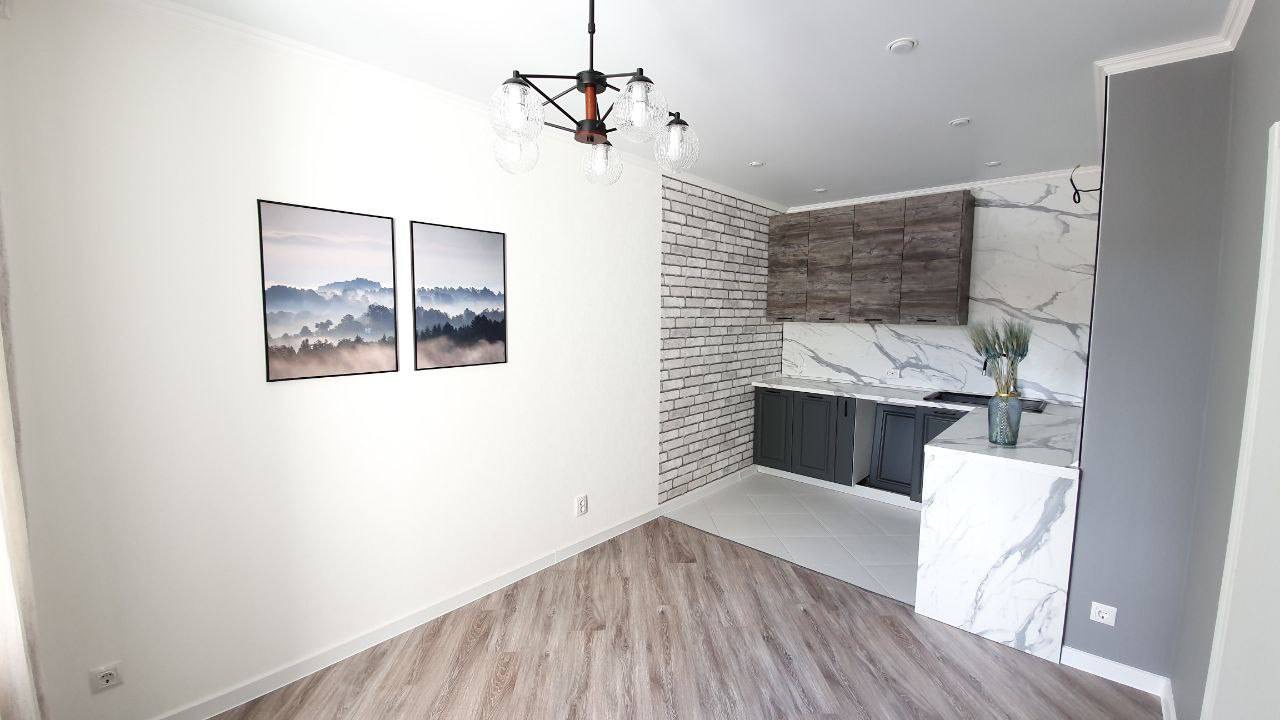 Nowoczesna kuchnia z marmurowym blatem, szarymi szafkami, ceglaną ścianą i drewnianą podłogą, oświetlona ozdobnym żyrandolem z kulistymi kloszami, udekorowana dwoma obrazami z motywem górskim.