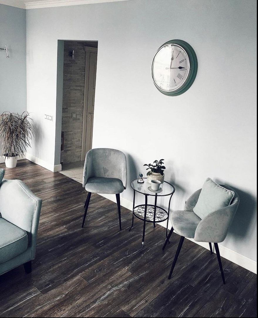 Jasne, pastelowe wnętrze z pomalowanymi ścianami, ciemną podłogą imitującą drewno, zegarem ściennym w zielonej ramie, dwoma fotelami i stolikiem kawowym z rośliną.