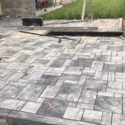 Fragment układanej nawierzchni z kostki brukowej w odcieniach szarości, widoczne narzędzia, poziomica, łata, fragmenty niezakończonej powierzchni, oznaczenie X na jednej z kostek.