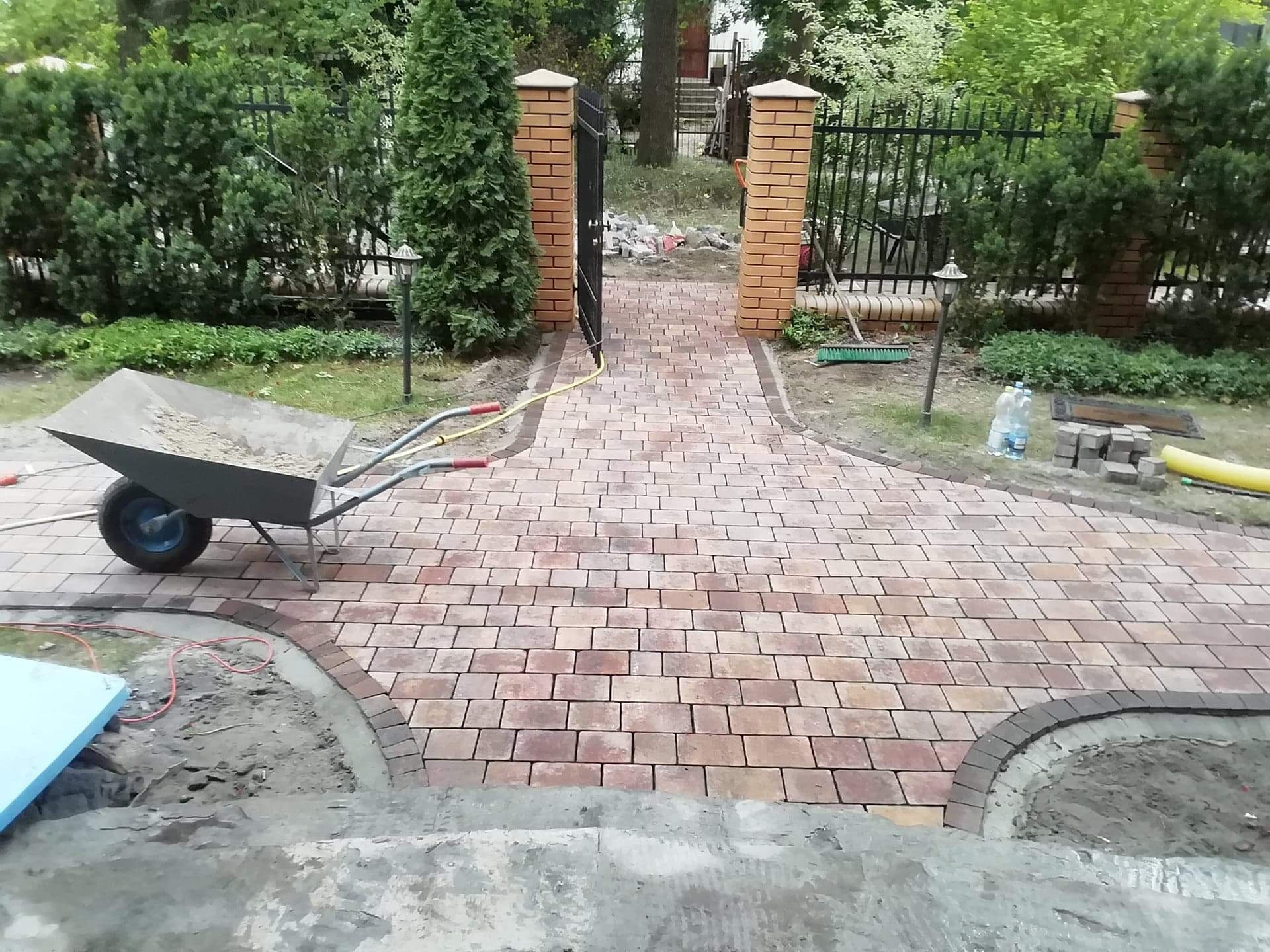 Ścieżka z kostki brukowej w trakcie układania, widoczny wózek z piaskiem, brama z ceglanymi słupkami i narzędzia ogrodnicze w tle.