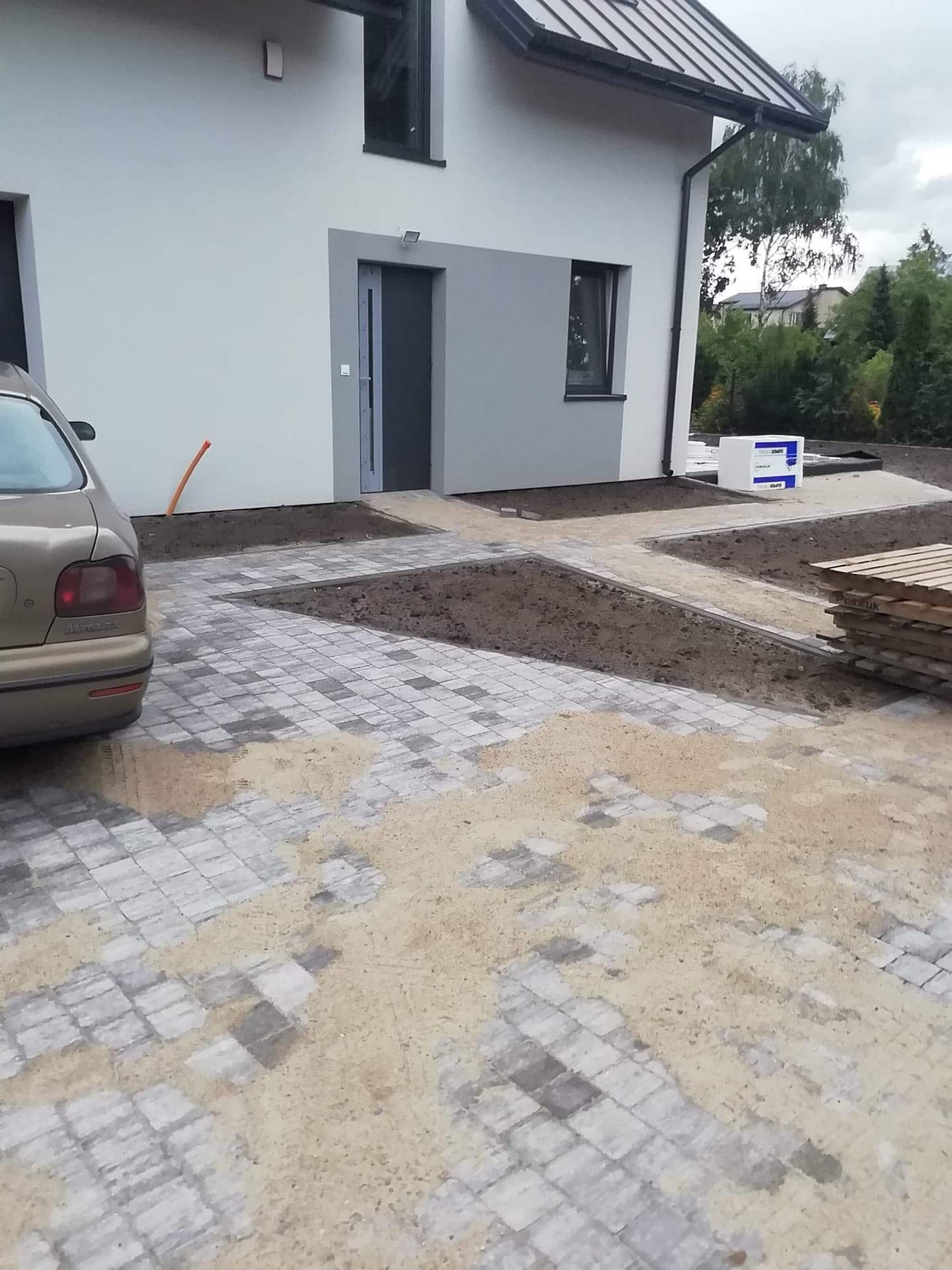 Fragment posesji z nową kostką brukową w odcieniach szarości, częściowo zasypaną piaskiem, widoczny samochód osobowy, przygotowane rabaty ziemne i paleta z kostką przy ścianie domu.