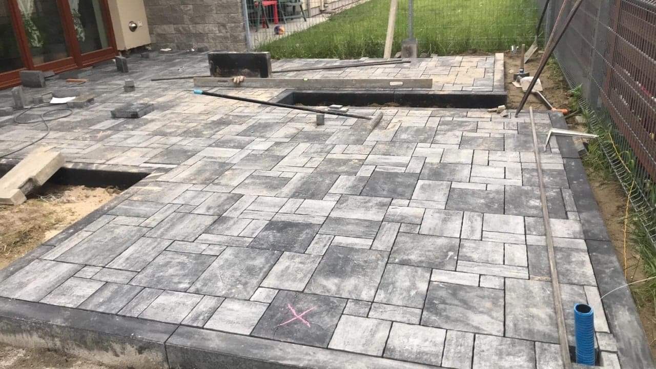 Fragment układanej nawierzchni z kostki brukowej w odcieniach szarości, widoczne narzędzia, poziomica, łata, fragmenty niezakończonej powierzchni, oznaczenie X na jednej z kostek.