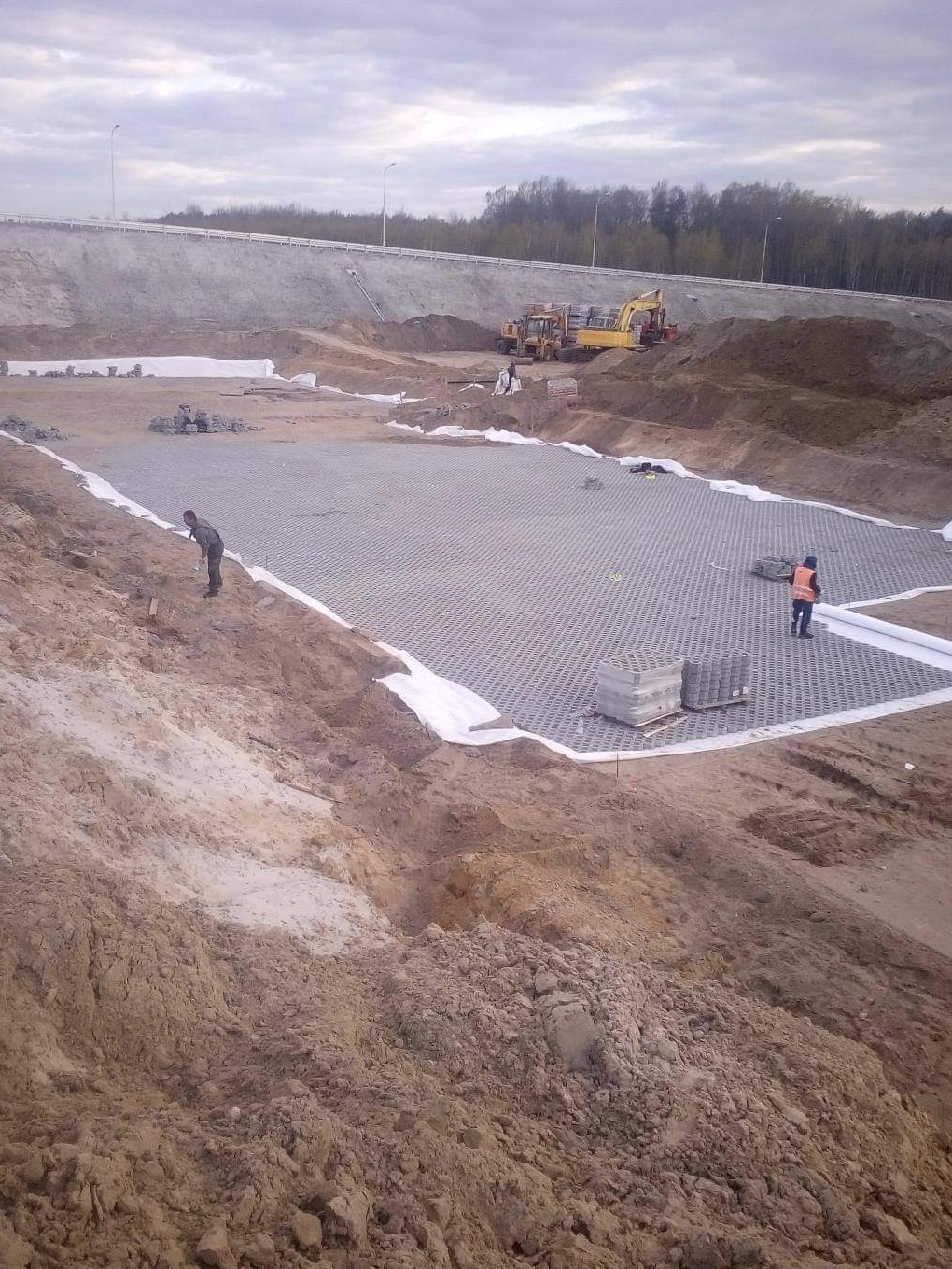 Przygotowanie podłoża pod kostkę brukową z widoczną geowłókniną, siatką wzmacniającą i ułożonymi kostkami. W tle koparka i dwaj pracownicy w kamizelkach odblaskowych.