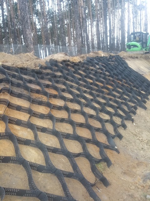 Siatka geokomórkowa umocniona na skarpie ziemnej, widoczny zielony traktor w tle, teren leśny.