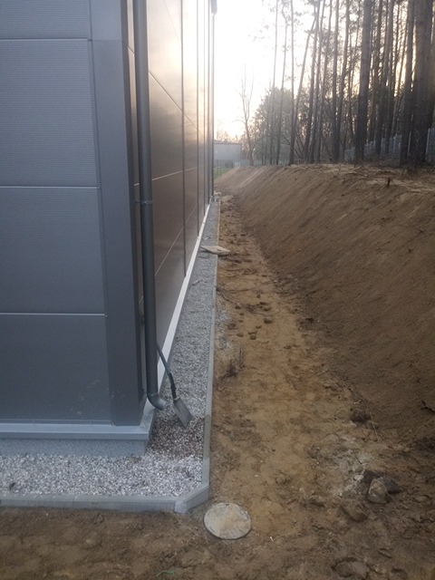 Szary budynek przemysłowy z widocznym systemem odwadniającym, obok wykopany rów z widoczną niwelacją terenu i przygotowaniem pod dalsze prace ziemne.