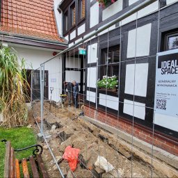 IdealSpace Sp. z o.o. - Prace ziemne przy zabytkowym budynku z muru pruskiego, widoczna wykopana ziemia, siatka zabezpieczająca, pracownicy używający ubijarki oraz tablica informacyjna firmy Ideal Space.