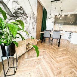 IdealSpace Sp. z o.o. - Nowoczesne wnętrze po remoncie: parkiet w jodełkę, designerska ściana z grafiką, drewniana komoda i zielony akcent roślinny. Kuchnia w tle, minimalistyczne lampy.
