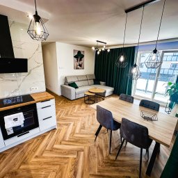 IdealSpace Sp. z o.o. - Nowoczesne wnętrze po remoncie: parkiet w jodełkę, szara sofa, stół z krzesłami i designerskie lampy wiszące. Widok na okno z zielonymi zasłonami.