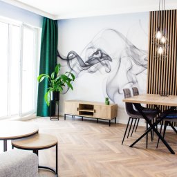 IdealSpace Sp. z o.o. - Jasne wnętrze po remoncie: parkiet w jodełkę, stół z krzesłami, sofa, szafka RTV, zielone zasłony i grafika na ścianie. Wykończenie pod klucz.