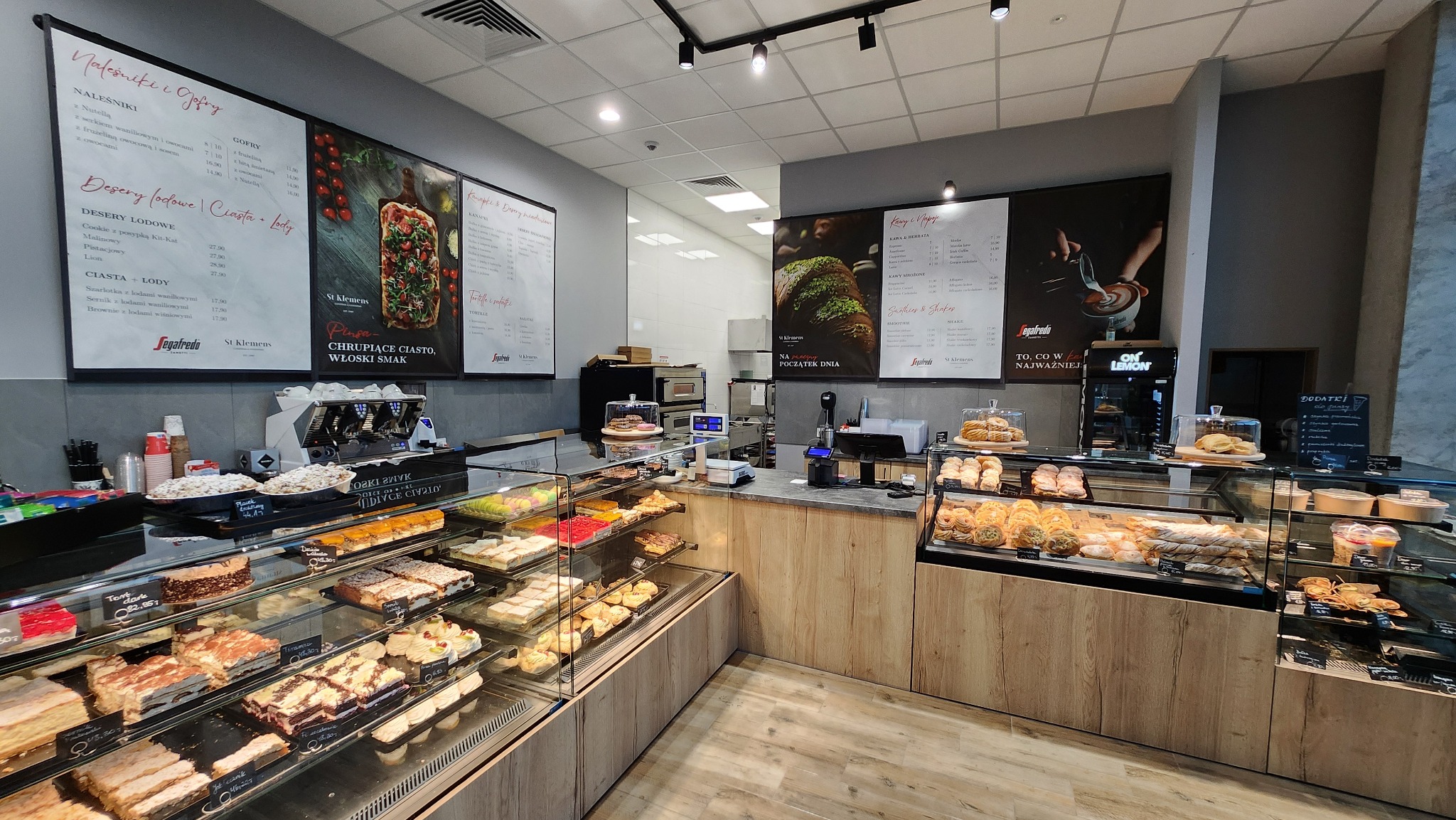 Wnętrze kawiarni St Klemens z przeszklonymi witrynami pełnymi ciast, deserów i pieczywa, menu na ścianach i ekspres do kawy w tle.