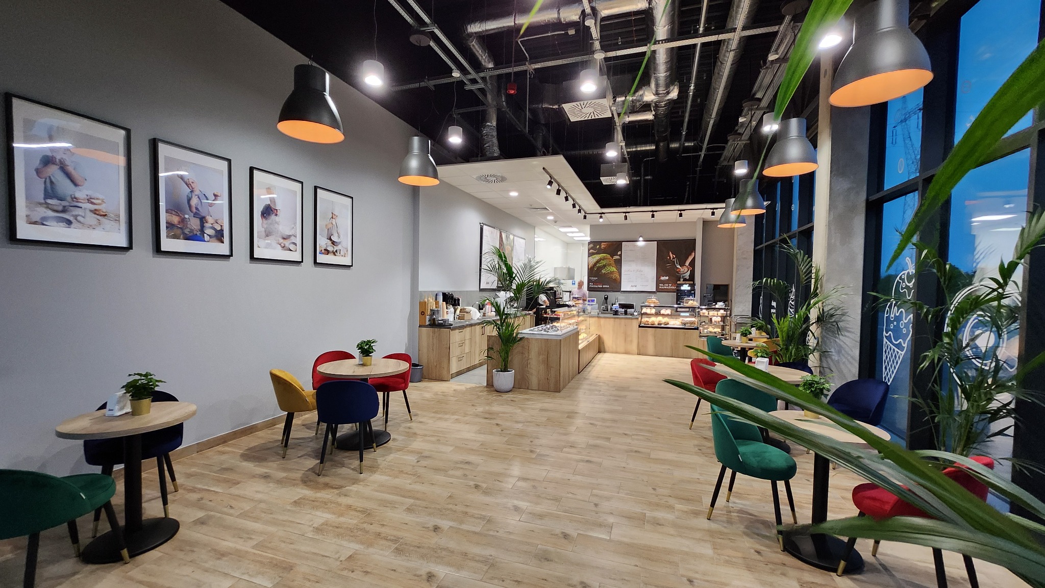 Wnętrze kawiarni z drewnianą podłogą, stolikami z kolorowymi krzesłami i roślinami doniczkowymi, oświetlone wiszącymi lampami, z widoczną ladą i ekspozycją ciast.