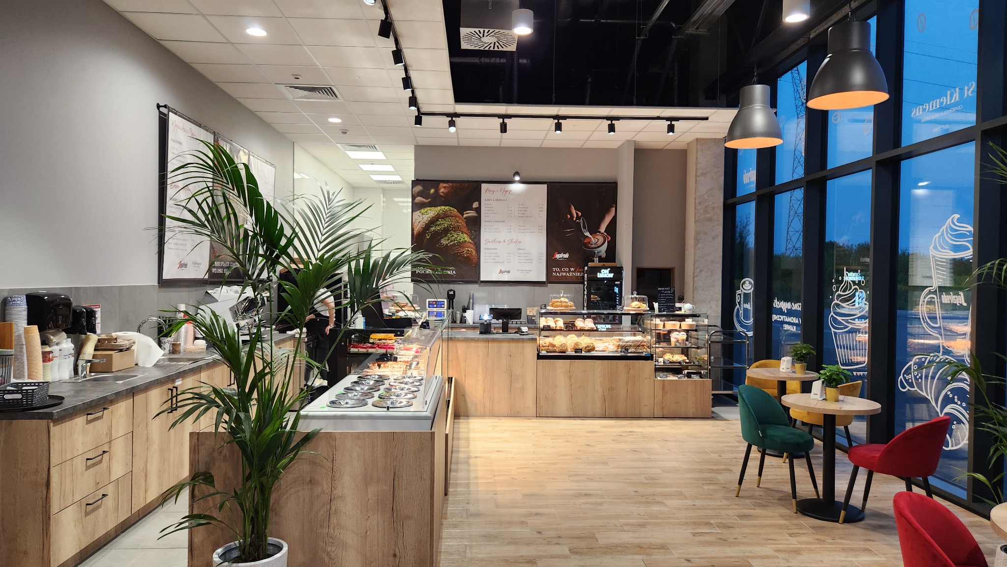 Wnętrze kawiarni St Klemens w Parku Handlowym BIG Gorzów: jasne drewniane podłogi, przeszklona witryna z ciastami, stoliki z kolorowymi krzesłami i duże okna z dekoracjami.