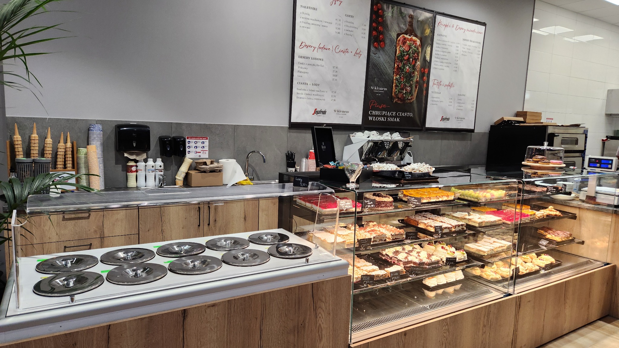 Wnętrze kawiarni: przeszklona lada chłodnicza z różnorodnymi ciastami i deserami, obok stanowisko do lodów z metalowymi pokrywami, w tle ekspres do kawy, menu na ścianie i zaplecze.