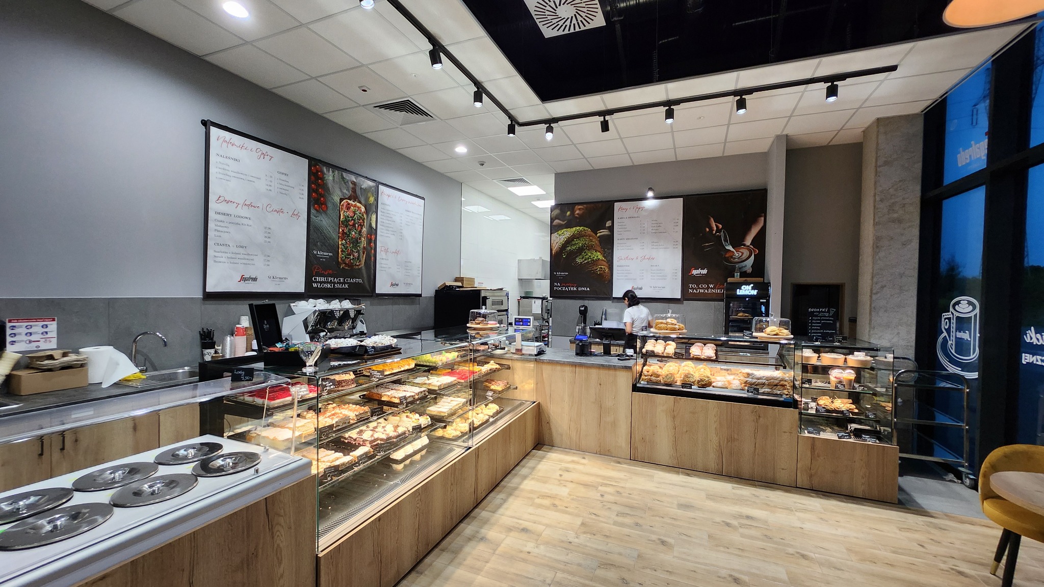 Wnętrze kawiarni St Klemens w Parku Handlowym BIG Gorzów z drewnianymi ladami chłodniczymi wypełnionymi ciastami i deserami, za którymi stoi pracownik w maseczce, a na ścianach wiszą menu z ofertą.