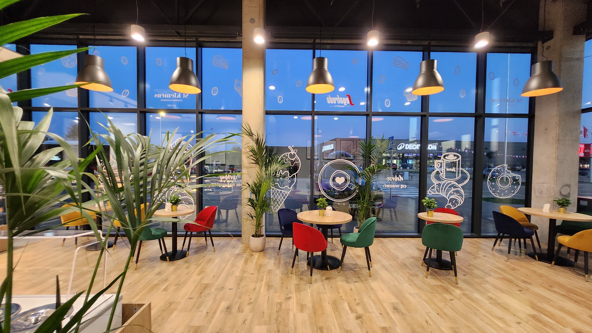 Wnętrze kawiarni St Klemens w Parku Handlowym BIG Gorzów z drewnianą podłogą, okrągłymi stolikami, kolorowymi krzesłami i dużymi oknami z naklejkami przedstawiającymi słodkości.