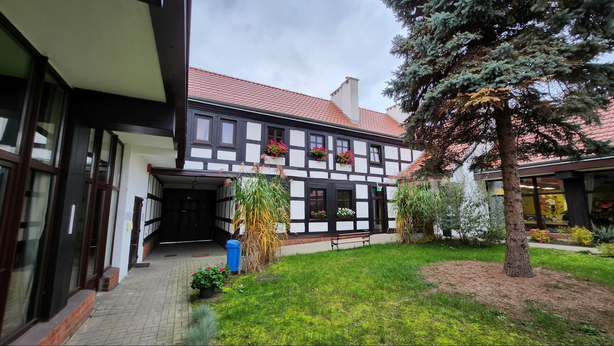 Budynek biblioteki publicznej w stylu szachulcowym po renowacji, z czerwonymi kwiatami w oknach i zadbanym trawnikiem przed wejściem.