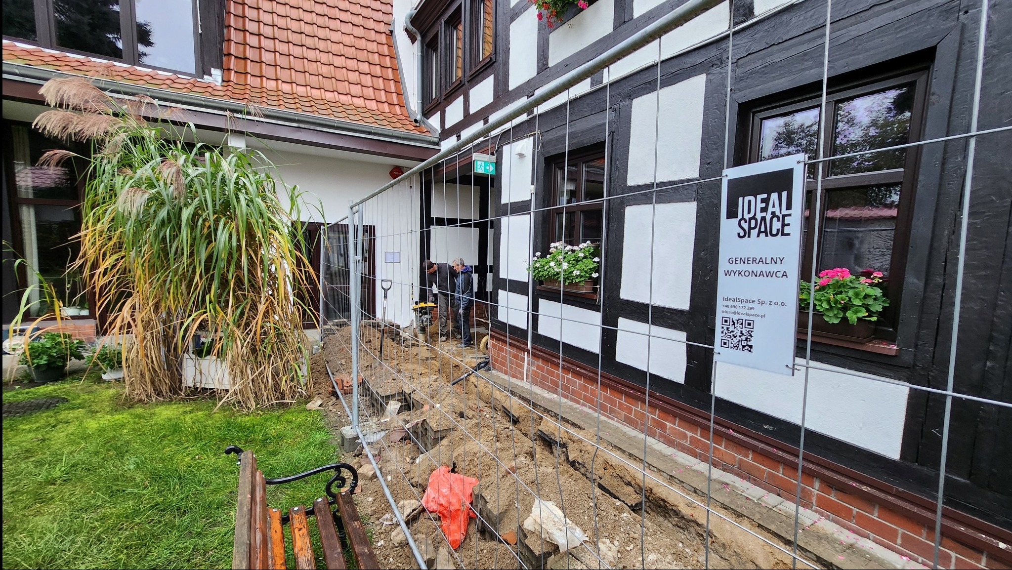 Prace ziemne przy zabytkowym budynku z muru pruskiego, widoczna wykopana ziemia, siatka zabezpieczająca, pracownicy używający ubijarki oraz tablica informacyjna firmy Ideal Space.