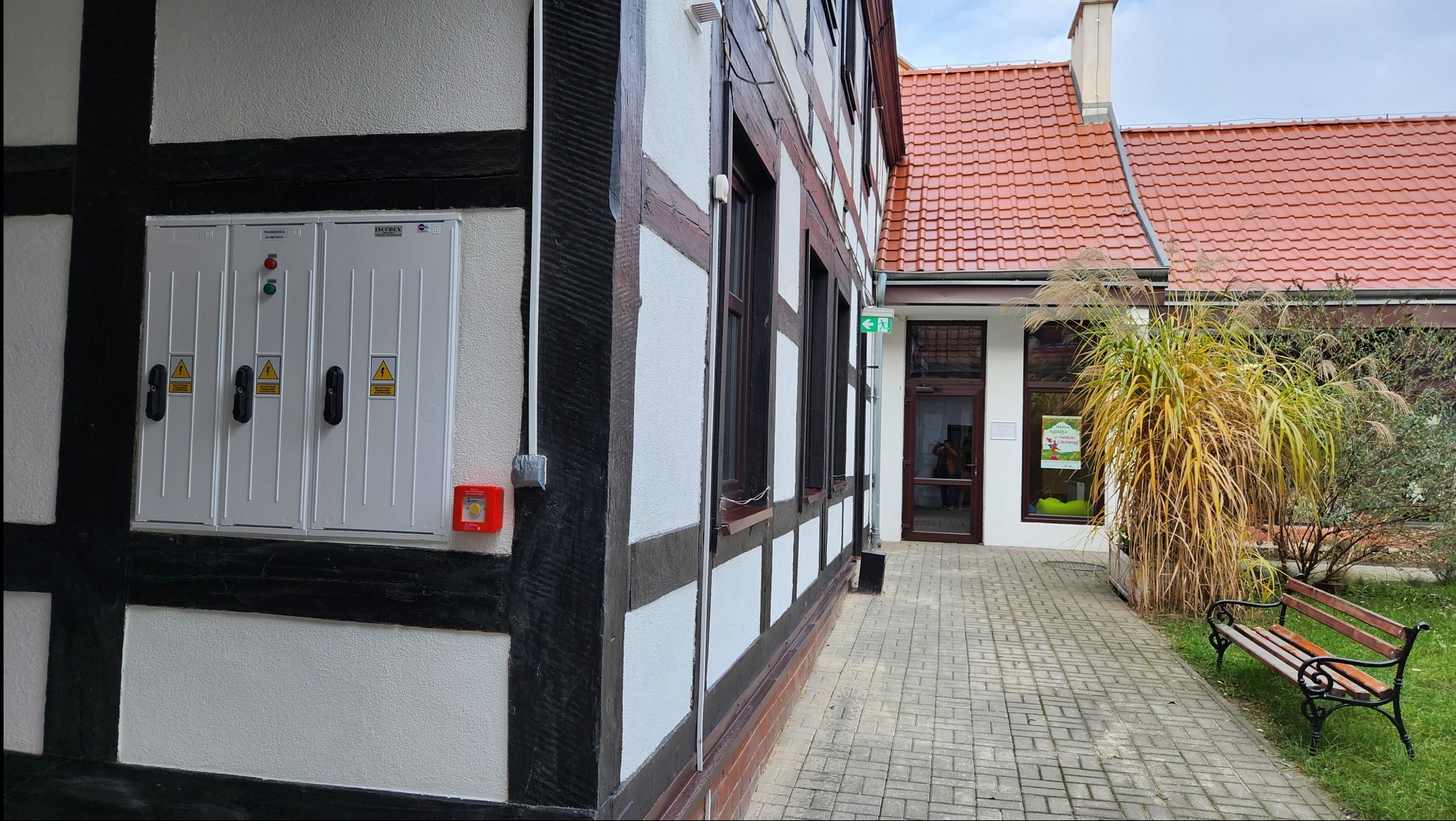 Biała skrzynka elektryczna z ostrzeżeniami na ścianie budynku biblioteki w stylu szachulcowym, obok czerwony alarm przeciwpożarowy, widok z zewnątrz na chodnik i wejście.