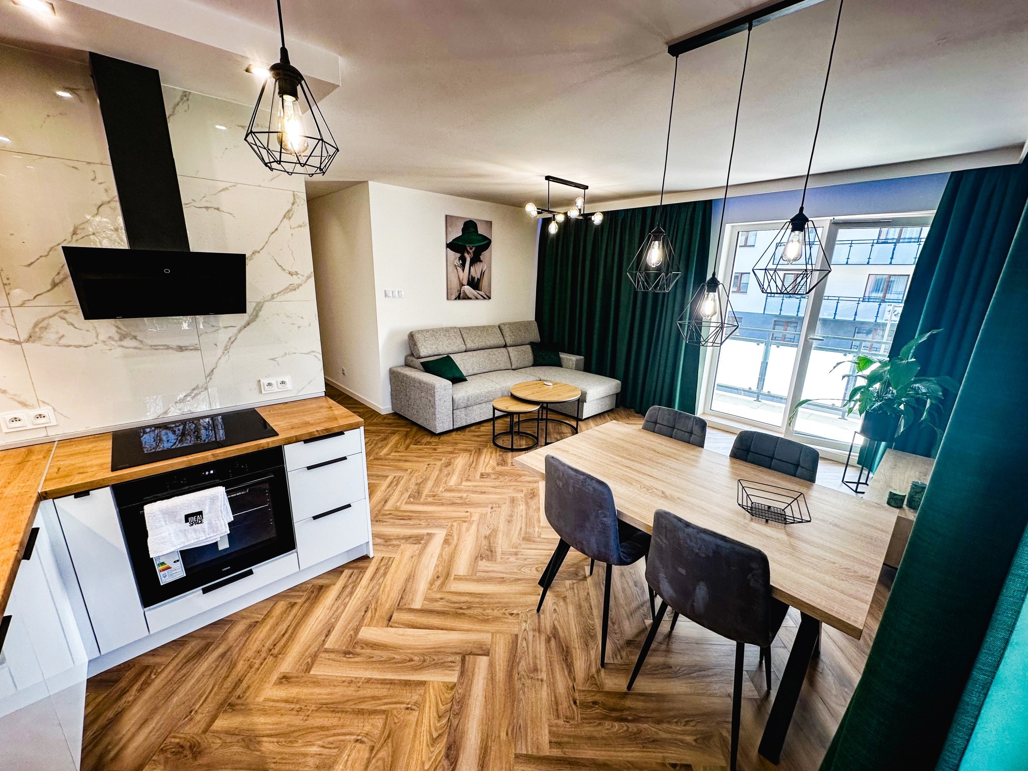 Nowoczesne wnętrze po remoncie: parkiet w jodełkę, szara sofa, stół z krzesłami i designerskie lampy wiszące. Widok na okno z zielonymi zasłonami.