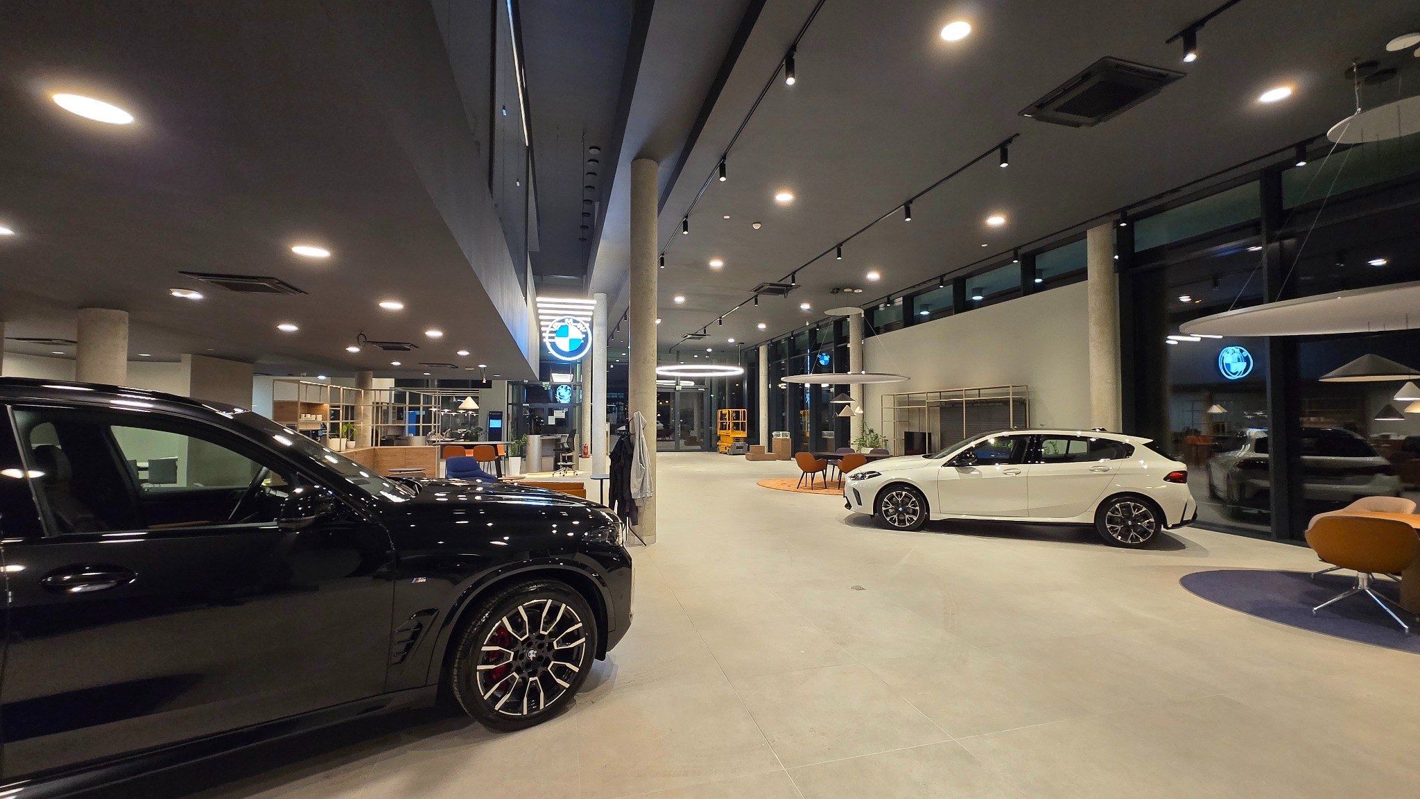 Wnętrze salonu samochodowego BMW po remoncie. Widoczne dwa samochody, czarny i biały, nowoczesne oświetlenie i przestronne wnętrze z elementami wystroju.