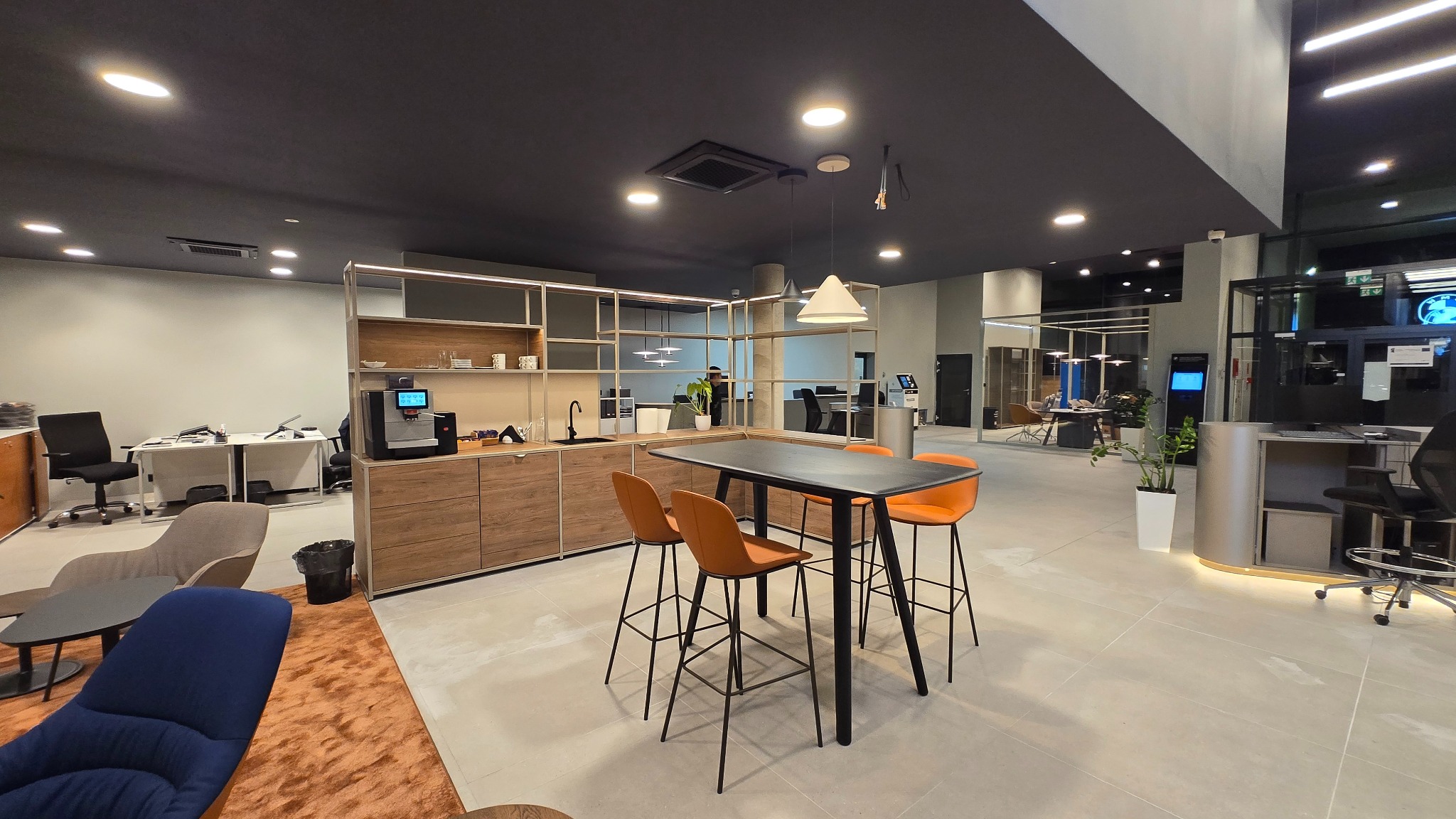 Nowoczesne wnętrze salonu samochodowego po remoncie: minimalistyczny design, czarne meble, pomarańczowe krzesła barowe, drewniane szafki, oświetlenie punktowe.