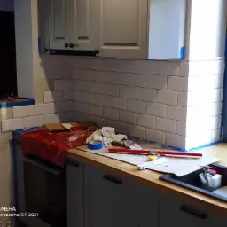 Białe płytki ułożone na ścianie w kuchni, widoczne narzędzia glazurnicze, poziomica, szpachelka, niebieska taśma malarska, resztki zaprawy i brud na blacie. W tle okno z widokiem na ogród.