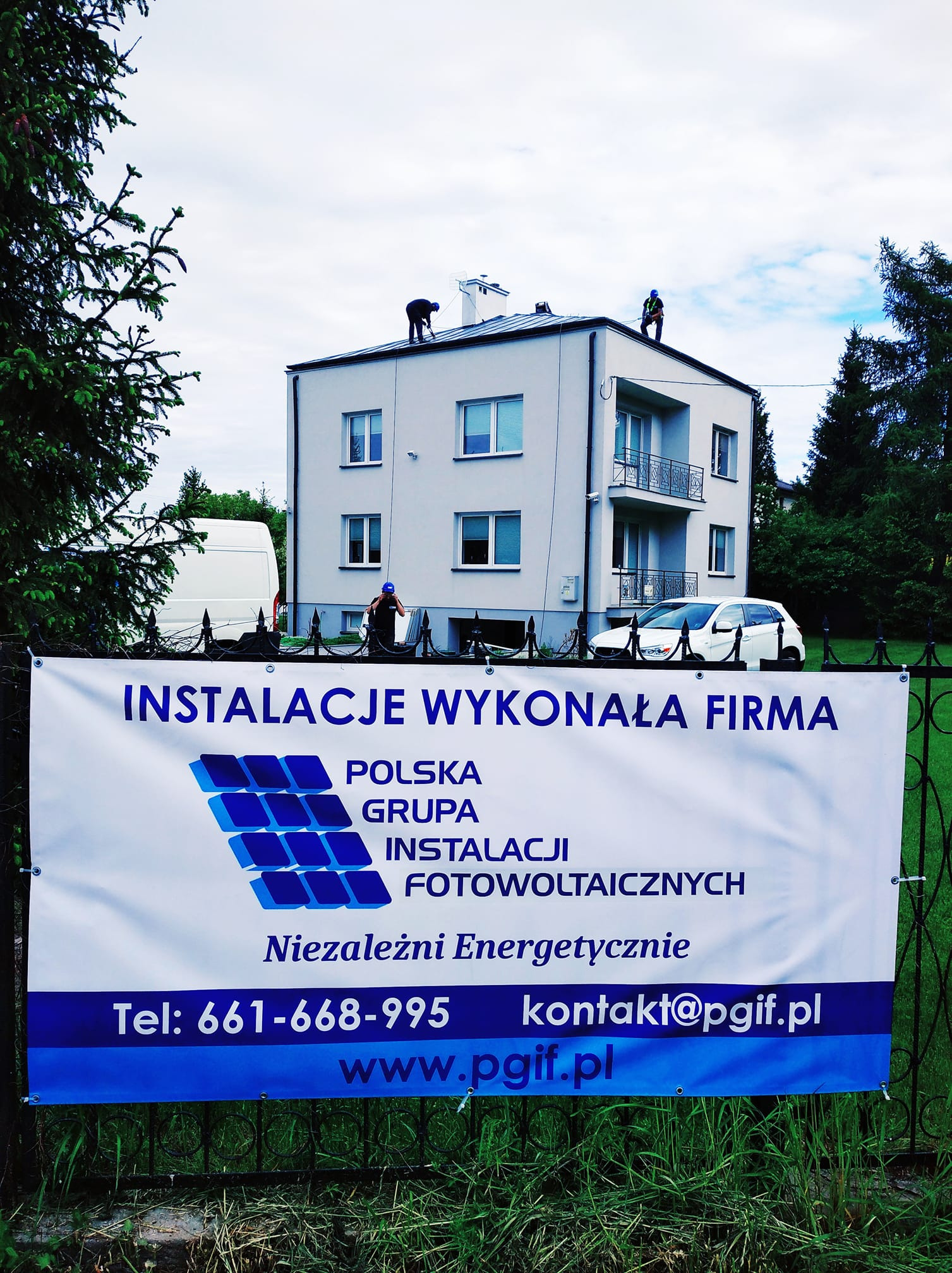 Montaż paneli słonecznych na dachu dwupiętrowego budynku, widoczny baner reklamowy firmy instalacyjnej z numerem telefonu i adresem e-mail, biały samochód i bus w tle.