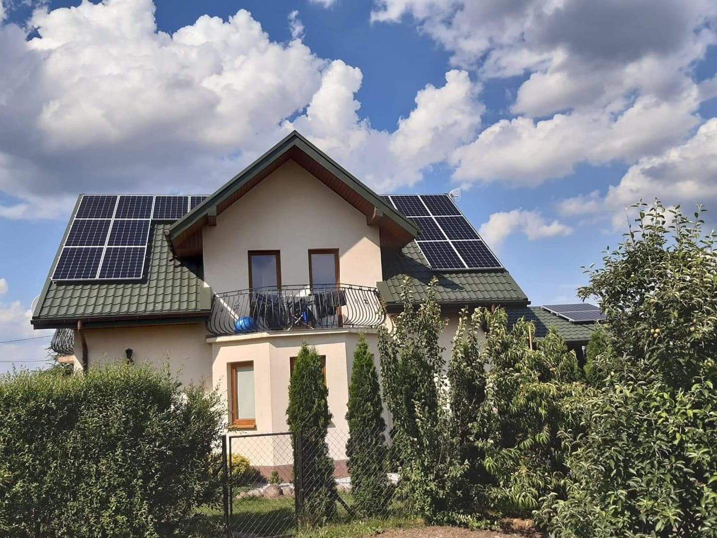 Dom jednorodzinny z panelami słonecznymi na dachu pokrytym zieloną dachówką, widok z poziomu gruntu, błękitne niebo z białymi chmurami w tle.