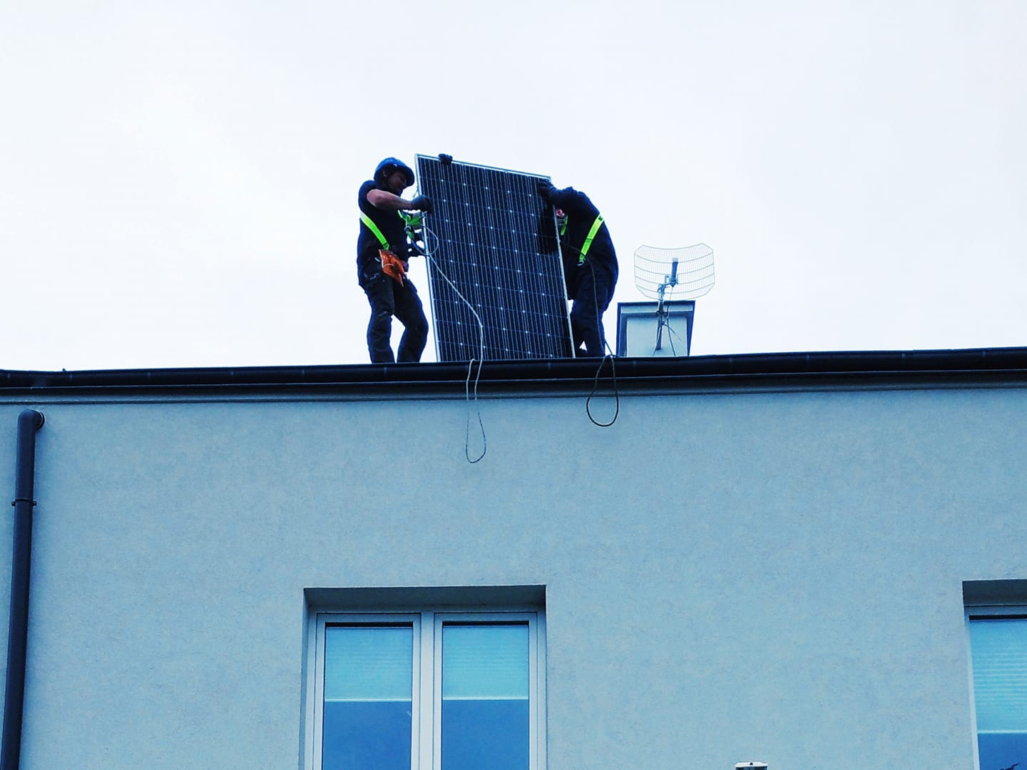 Dwóch instalatorów w uprzężach na dachu budynku montuje panel słoneczny, widoczna antena telewizyjna w tle.