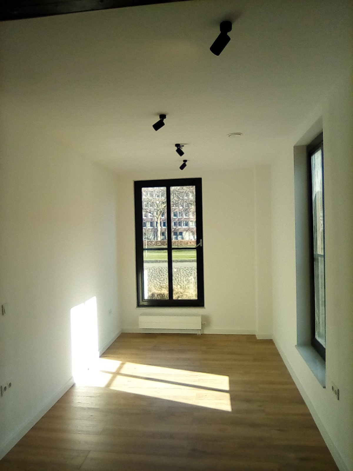 Puste, wykończone pomieszczenie z drewnianą podłogą, białymi ścianami, czarnymi ramami okiennymi i reflektorami punktowymi na suficie, wpadające promienie słoneczne.