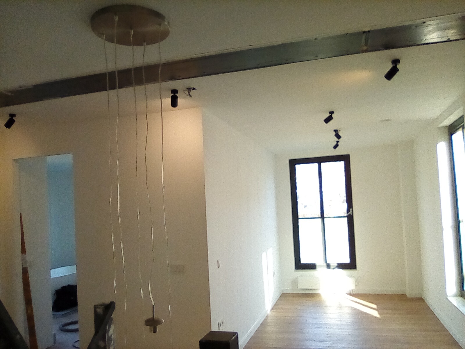 Jasne wnętrze z drewnianą podłogą, nowoczesnym oświetleniem punktowym i wiszącą lampą z wieloma kablami, widok na okno z czarną ramą.