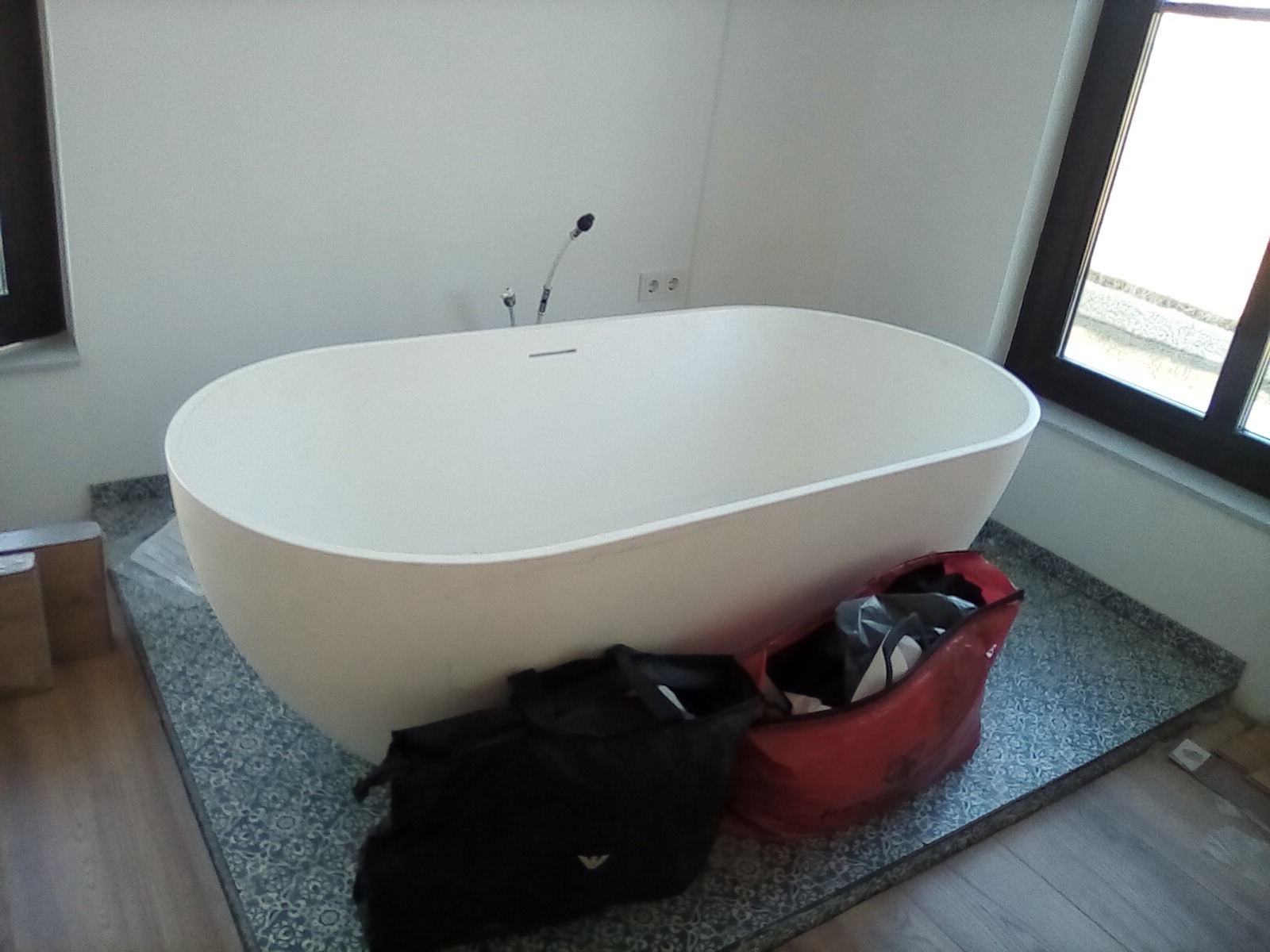 Biała, owalna wanna wolnostojąca na tle ściany z oknem, z widocznymi torbami budowlanymi i niedokończoną podłogą, sugerująca etap prac wykończeniowych w łazience.