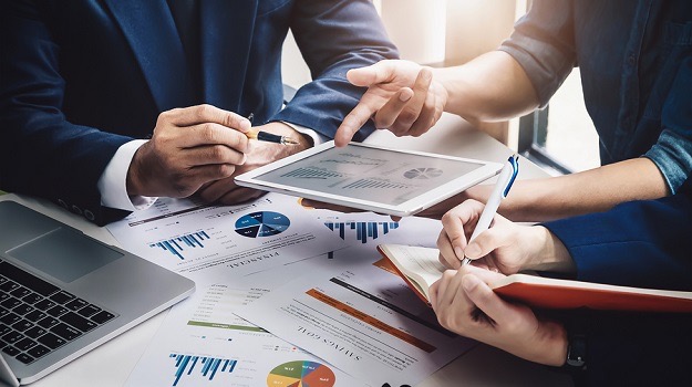 Zespół analizuje dane finansowe na tablecie i w notatniku, otoczeni wykresami i laptopem.